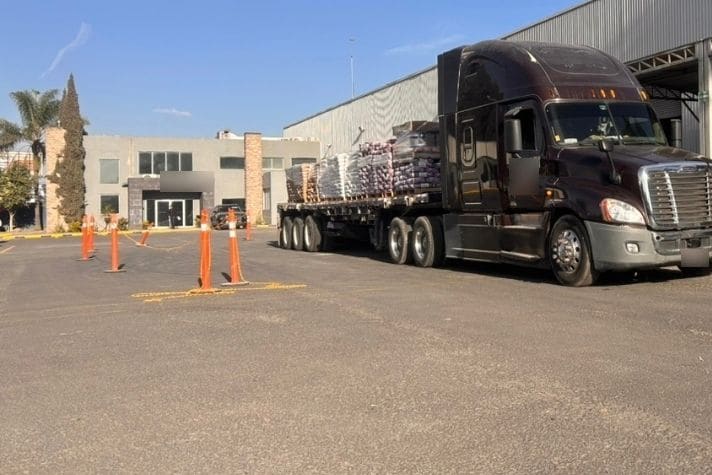 Tractocamión con plataforma cargada de mercancía paletizada en el patio de una planta industrial.